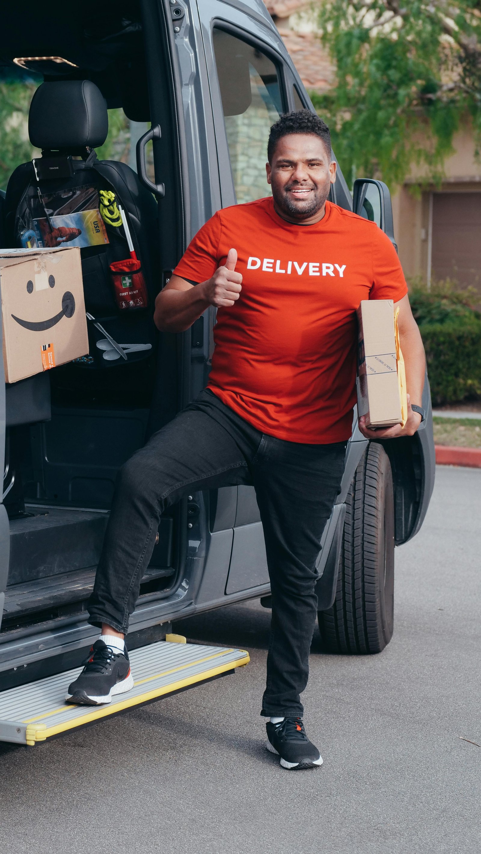 Cheerful delivery man in a red shirt holding a package and giving a thumbs up by his van.